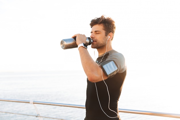 Uomo sportivo in maglietta ascoltando musica tramite cuffie wireless e acqua potabile dalla tazza di metallo, dopo l'allenamento in riva al mare