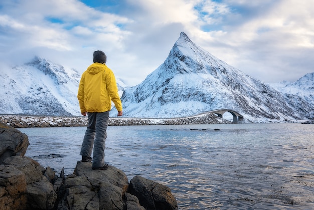 Спортивный человек в желтой куртке, стоя на камне на берегу моря против снежных гор и облачного неба на закате зимой