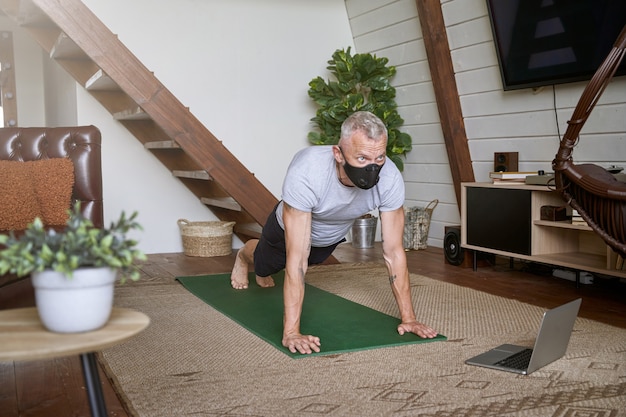 Sporty middle aged man wearing face protective mask standing in plank pose while exercising at home