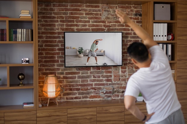 Photo sporty middle aged man working out at home watching sport channel and fitness workout program on tv back view