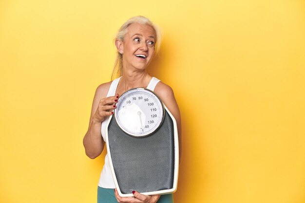 Photo sporty middleaged caucasian woman holding scale yellow studio looks aside smiling cheerful