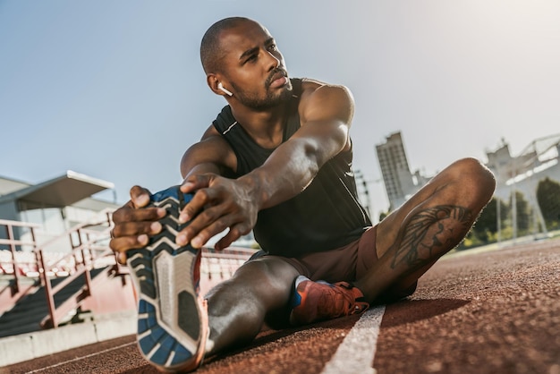 Sporty muscular african male athlete in earphones looking away stretching legs while sitting at the stadium race track