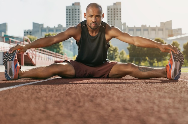 Sporty muscular african male athlete in earphones stretching legs while sitting at the stadium race