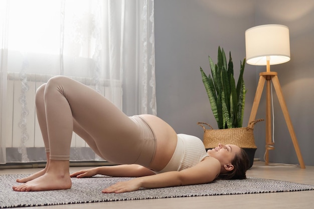 Sporty pregnant woman wearing beige top and leggings doing bridge exercise training her body taking care of health during pregnancy enjoying home gymnastic