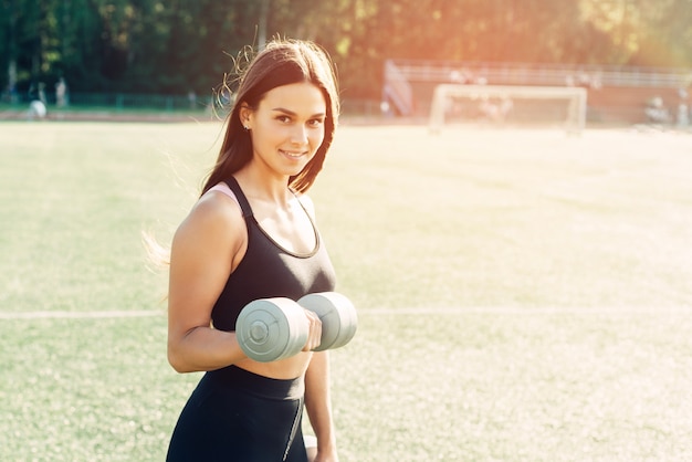 Спортивная улыбающаяся девушка с гантелями в руках на стадионе. Здоровый образ жизни