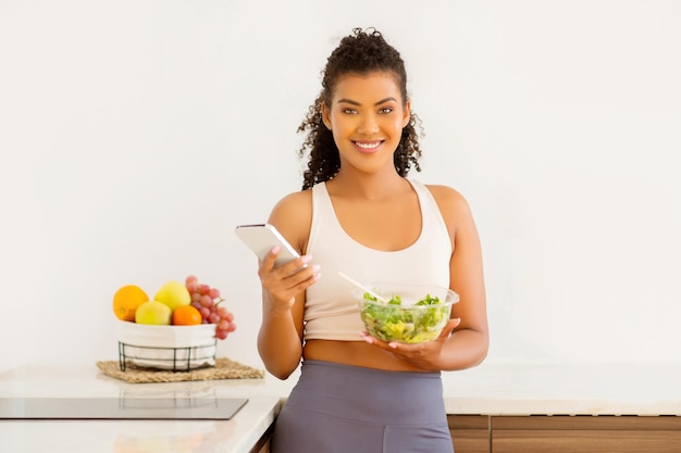 Sporty vrouw gebruikt telefoon poseert met salade schaal in de keuken