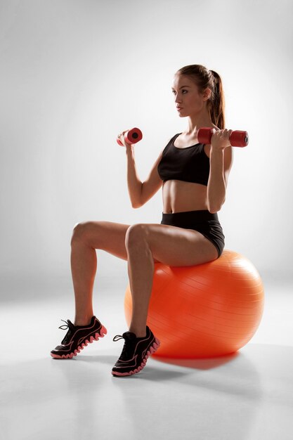 Sporty woman doing aerobic exercise with red dumbbells on a fitness ball on grey wall