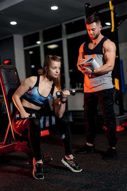 Sporty woman doing sport with trainer. Fitness muscular man writing in notebook. Pretty female working hard in gym.