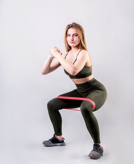 Sporty woman doing squats using rubber resistance band isolated on gray background