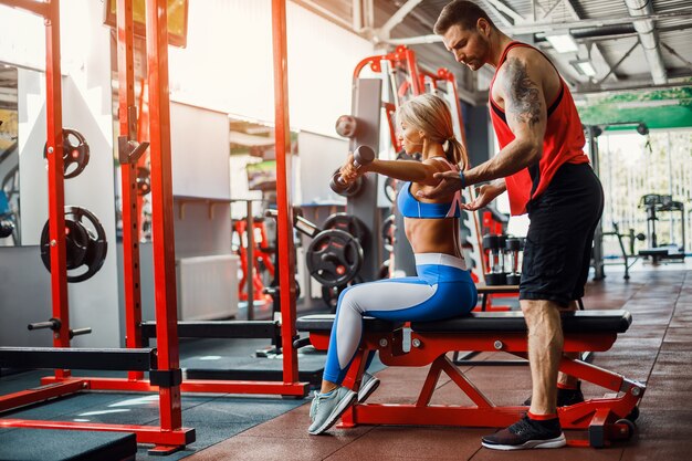 Sporty woman doing weight exercises with assistance of her personal trainer at gym