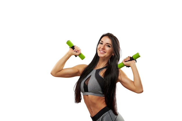 Спортивная женщина в серой спортивной одежде позирует и улыбается в камеру, накачивая мышцы плеч и бицепсы. Отдельный на белом фоне.