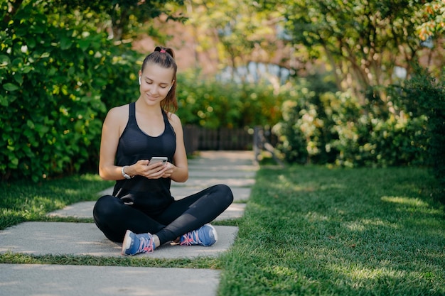 La donna sportiva ha una pausa per il ristoro controlla i messaggi e la notifica condivide i contenuti sul fitness all'aperto vestita con abbigliamento attivo conduce uno stile di vita sano si rilassa dopo l'allenamento cardio attivo