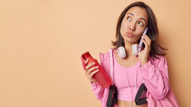 Sporty woman has thoughtful expression has telephone\
conversation carries sport equioment holds bottle of water rests\
after workout isolated over beige background empty space for\
advertisement