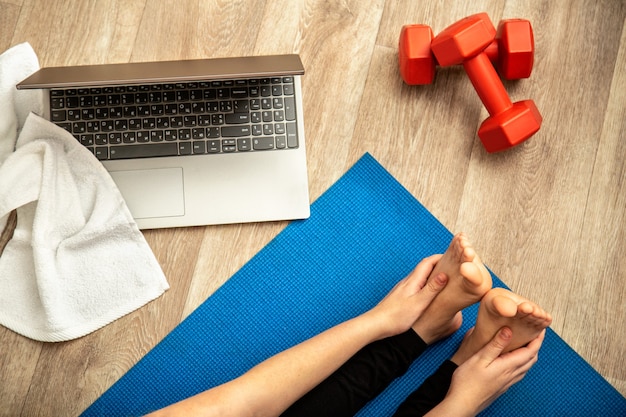 Sporty woman making yoga fitness stretching at home via laptop. Young woman losing weight by online gym workout using dumbbells on house floor top view. Body health and wellness.