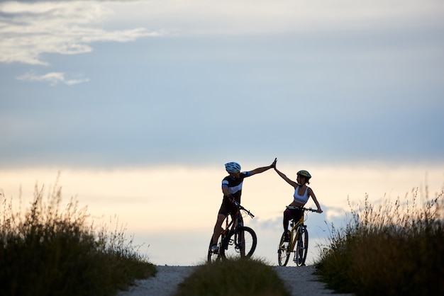 Sporty woman and man riding bicycles, having fun outside. Silhouettes of sportsmen highing five and posing on road in sunset time. Non urban scene.