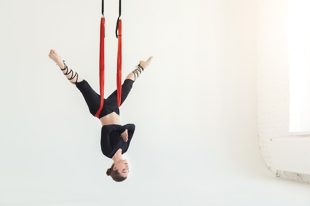 Sporty woman practicing fly yoga asana, hanging head down over white background in fitness gym, copy space. Health, sport, yoga concept