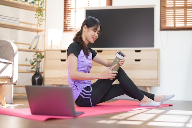 Sporty woman sitting on yoga mat to drinking protein shake after doing exercise yoga at home