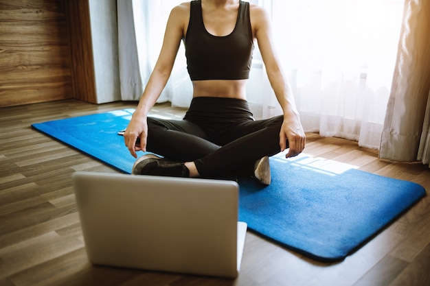 A sporty woman in sportswear is sitting on the floor with dumbbells and a protein shake or a bottle of water and is using a laptop. Sport and recreation concept.