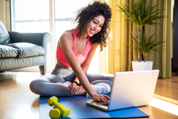 Sporty woman in sportswear is sitting on the floor with dumbbells using a pc laptop in the living room