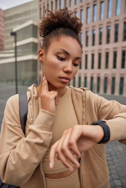 Sporty woman with curly hair tracks pulse concentrated into smartwatch keeps hand on neck monitors progress dressed in sportswear poses against building. Fitness lifestyle and health concept