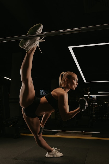 A sporty woman with long blonde hair is doing a twine during dumbbell bicep curls in a gym. A girl is stretching.