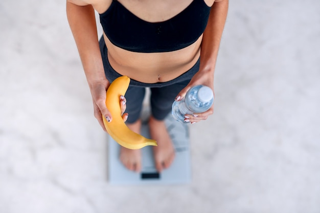 Sporty woman with a perfect body measuring body weight on electronic scales and holding a yellow banana and a bottle of water