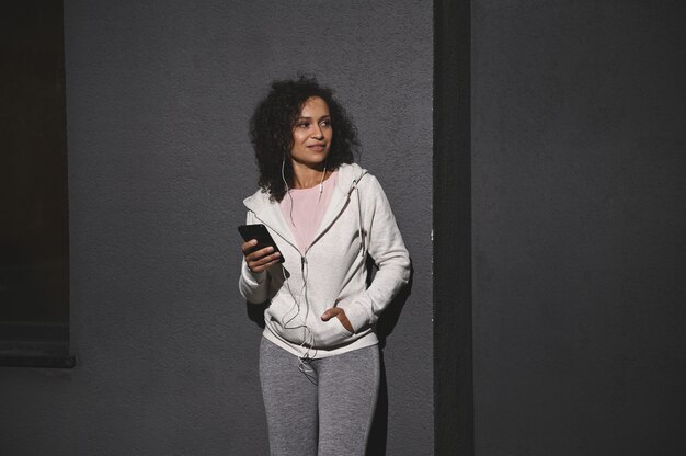 Sporty young African woman listening to music on earphones, holding a mobile phone relaxing after workout, leaning on a grey wall of urban building