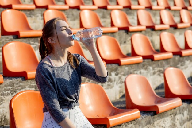 La giovane ragazza attraente sportiva in abiti sportivi che si rilassano dopo l'allenamento duro si siede e beve l'acqua dalla bottiglia di sport speciale dopo avere corso