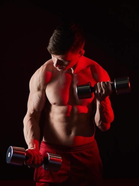 Sporty young man training with dumbbells against dark background