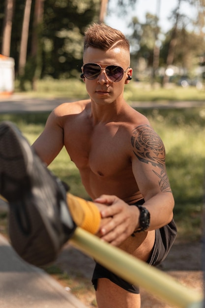 Sporty young man with hairstyle sunglasses and headphones in black sneakers and shorts doing stretching and workout in the park