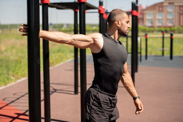 Sporty young muscular man in black vest and shorts holding by sports bar while exercising outdoors