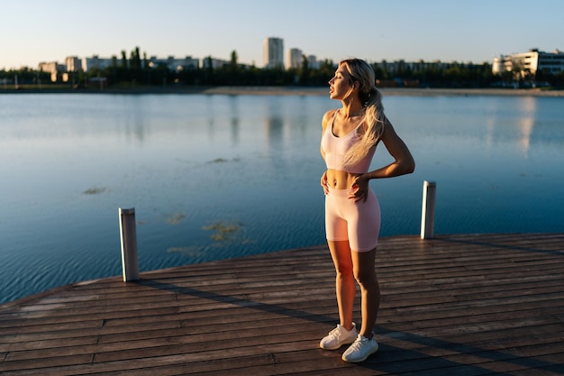 Sporty young woman exercising and stretching neck for warming up before running or working out near water on background of cityscape