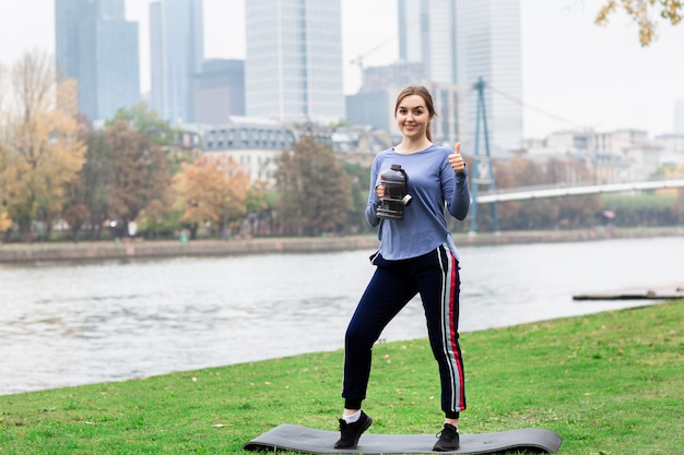 Sporty young woman posing in the city near the river. A woman holds a water bottle in one hand and shows a class in the other