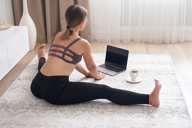 La giovane donna sportiva si siede sul pavimento nel soggiorno pratica lo yoga sulla lezione di computer online femminile