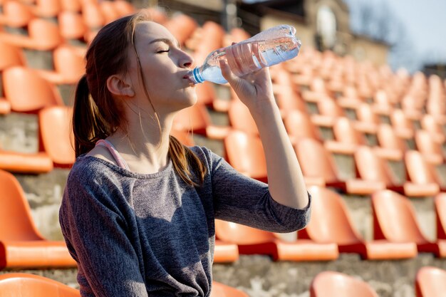 La giovane donna sportiva in abiti sportivi che si rilassano dopo l'allenamento duro si siede e beve l'acqua dalla bottiglia di sport speciale dopo avere corso su uno stadio