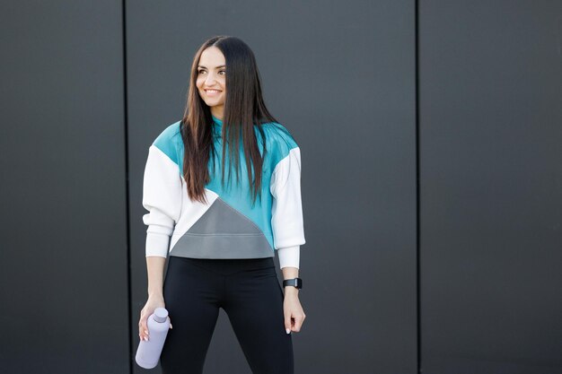 Sporty young woman standing and holds bottle with fresh water dressed in activewear poses outdoors Healthy lifestyle