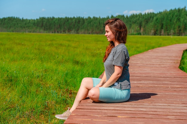 A sporty young woman walks along a picturesque wooden walking path through a swamp with tall grass