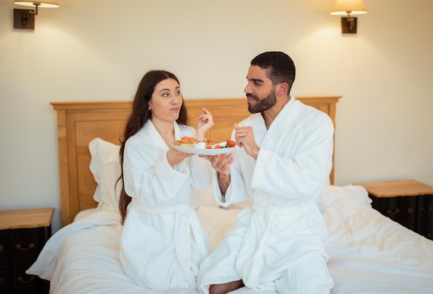 Spouses eating breakfast in bed wearing white bathrobes indoors