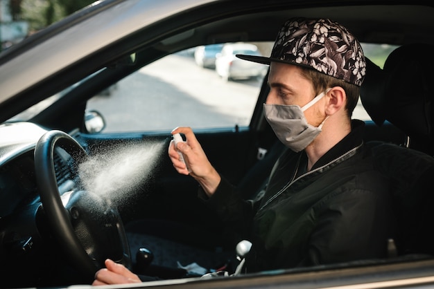 Spraying antibacterial sanitizer spray on steering wheel, disinfection car, infection control concept. Prevent Coronavirus, COVID-19, flu. Man wearing in medical protective mask driving a car.