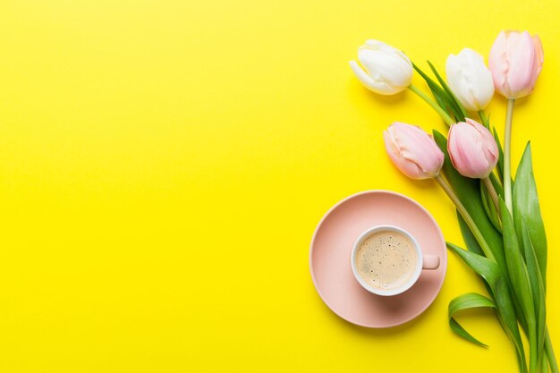 春の背景に花、一杯のコーヒー、テキスト用の場所を持つ色付きのテーブル背景にピンクと白のチューリップの花束 コピースペーストップビュー