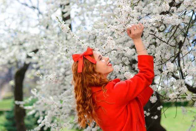 Spring beauty girl with long red blowing hair outdoors. Blooming trees. Romantic young woman portrait. Nature