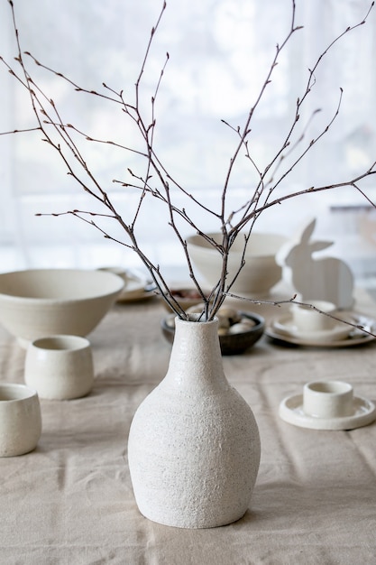 Spring birch branches in white ceramic vase standing on table with linen tablecloth and Easter decorations.