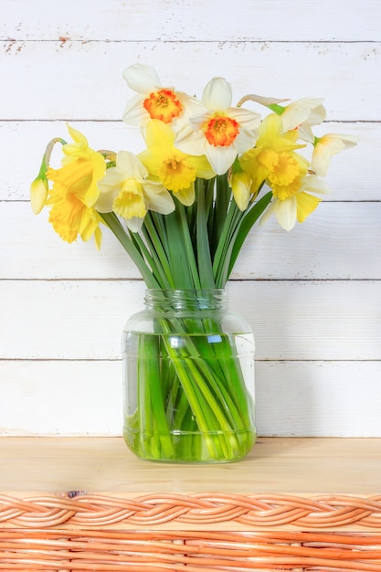 Spring bouquet of Narcissus flowers in a glass jar on a white wooden wall close-up