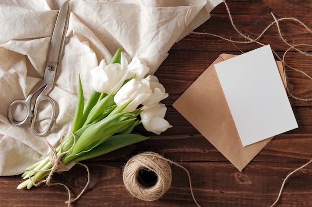 白いチューリップの花、空白の紙カード、はさみ、素朴な木製の机の上のひもの春の花束。
