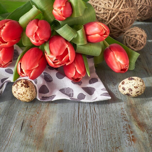 Весенний фон празднования: букет красных тюльпанов, весенние украшения и крашеные яйца на серой деревенской доске.
