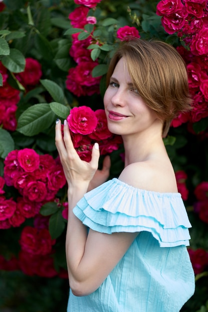 春。庭に咲くバラの臭いがするスタイリッシュな青い光のドレスを着ている肖像画魅力的な赤毛の若い女性を閉じます。アウトドア。