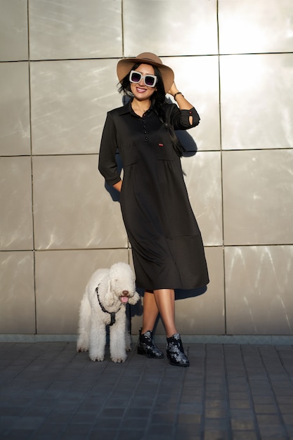 봄 컬렉션. 세련 된 검은 드레스와 귀여운 강아지와 함께 포즈 유행 선글라스를 착용 행복 예쁜 갈색 머리 소녀. 회색 벽 배경에 포즈 유행 복장 모델. 야외 초상화.