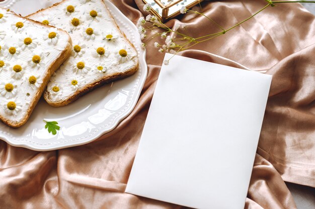 Весенняя композиция, белый чистый лист бумаги, бутерброд из тостового хлеба с цветами ромашки в белой тарелке
