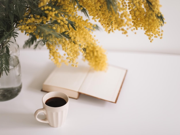 コーヒーカップの本と白いテーブルの背景に黄色のミモザの花と春の構成