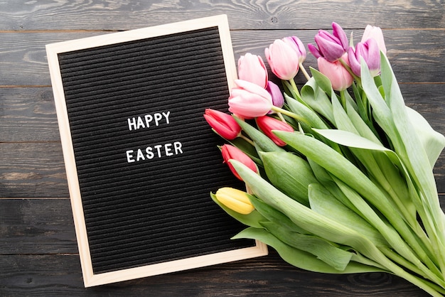 Spring concept. Bouquet of colorful tulip flowers and letter board with the words Happy Easter top view flat lay on black wooden background